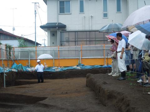 発掘現場と思われる場所で、白いヘルメットをかぶった説明者が傘を差しながら立ち、雨の中で見学者たちが傘を持って発掘調査の説明を熱心に聞いている様子の写真