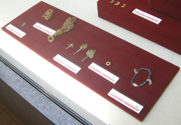 赤色の敷物の上に複数の展示品が点々と並び、それぞれに白い説明書きが添えられている、古墳から出土したと見られる副葬品の展示写真