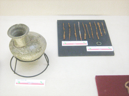 平たい台の上に土器のような丸い壺と複数の細長い金属片が展示され、それぞれに白い説明書きが添えられている博物館の展示写真
