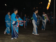 上に吊るされた提灯の明かりに照らされた夜の屋外会場で、青い浴衣を着た複数の女性たちが、輪になって「ささら舟」をおどっている様子の写真