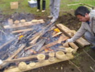 火を焚いて土器を焼く屋外の作業現場で、木炭と燃えさしの中で土器が焼かれており、作業服の男性が棒を使い土器の様子を確認している、土器製作の様子を捉えた写真