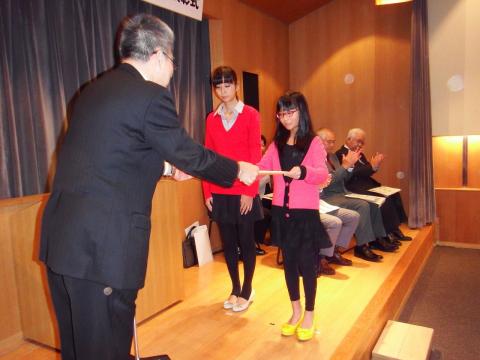 ステージ上でスーツ姿の男性が対面に並んで立った2人の女性に表彰状を手渡している様子の写真