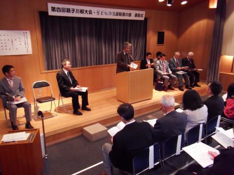 ステージ上で、スーツ姿の男性たちが左右に並んだ椅子に座る中、中央の演台に立った男性が、客席の出席者たちに向かって話をしている様子の写真