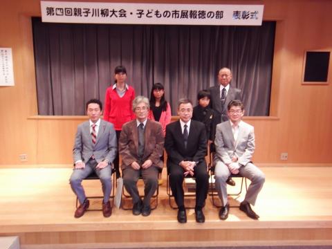 第四回親子川柳大会と書かれた横断幕が掲げられたステージの上で、6人のスーツ姿の男性と女性が椅子に座り、その後ろに5組の親子が並んで立って写した記念写真