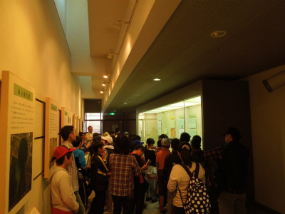 屋内通路の片側に展示パネルが並び、もう一方の側にはガラスケースの展示がある展示館で、たくさんの子供たちが熱心に展示を見ている様子の写真