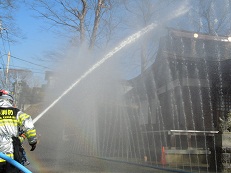 建造物の手前の地面に設置したホースから、幾筋も噴水上に水が噴き出し、その前では消防士が建物にめがけてホースから勢いよく水を噴射している消火訓練の様子の写真