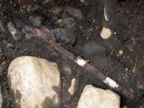 暗く湿った土の中に、錆びて茶色に変色した細長い金属の遺物が、周囲の石とともに出土した状態を接写した発掘現場の写真