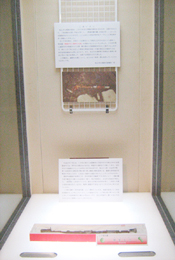 細長い銀装圭頭大刀が白い台の上に置かれ、その上には白いパネルと写真が展示されている博物館の展示ケースの写真