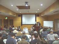 多数の参加者たちが椅子に座り、壇上の戸田哲也さんがスクリーンを指しながら説明している講演会の様子の写真