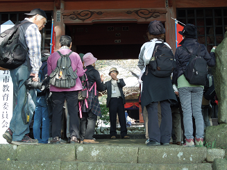 お寺の入り口の場所で、帽子をかぶった男性が歴史を語り、その周囲に集まったリュックサックを背負ったたくさんの人々が熱心に話を聞いている様子の写真