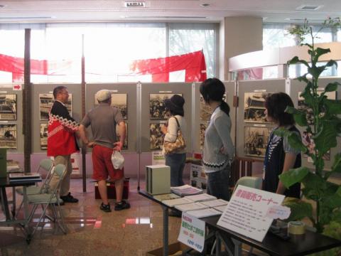 たくさんの白黒写真が掲示された展示パネルの前で男性と女性が法被を着た男性の説明を受けながら写真を見ており、手前にある書籍販売コーナーでは、長机に複数の冊子が並べられ係の女性2人が立っている様子の写真