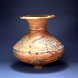 破損した赤褐色の土器の壺で、底部が三つの足に分かれ、表面には波打つ線や横向きの文様が刻まれており、口の部分がわずかに欠けている弥生前期壺形土器の写真