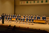 舞台上で、紺色の制服を着た大勢の保育園児が、指揮者に合わせて鉄琴や木琴といった打楽器を演奏している様子の写真