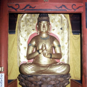 金色の布を背景に、豪華な赤い台座に座り、両手を合わせ、背景には金色の飾り板がある仏像が安置されている写真