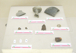 展示台の上に様々な形の土器や鉄器のような複数の遺物が並べられ、それぞれに白い説明書きが添えられている、博物館の展示品の俯瞰写真