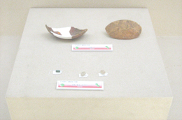 四角い台の上に割れた器や丸い石のようなものが並べられ、それぞれに白い説明書きが添えられている、考古学的な遺物の展示写真