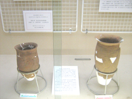 茶色の2つの壺が台の上に置かれ、白い修復跡が目立ち、それぞれに説明書きが添えられている、考古学的な遺物の展示写真