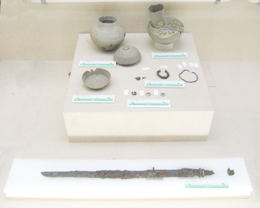 台座の上に壺や装飾品、皿などの遺物が展示され、その手前には錆びた刀剣のような細長い金属片が配置されている、考古学的な遺物の展示写真