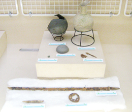 白色の台の上に壺が並べられ、台の下には長い金属片や丸い装飾品が展示されている、考古学的な遺物の展示写真