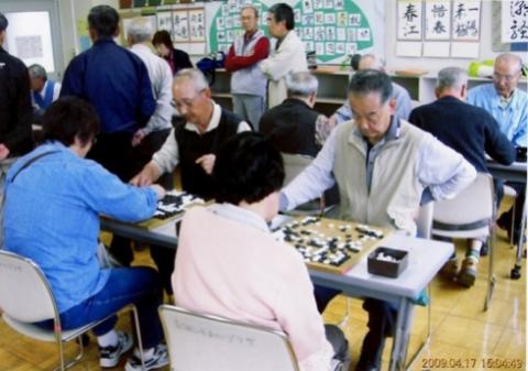碁石が置かれたテーブルを囲み、たくさんの高齢者の方々が囲碁を楽しんでいる写真