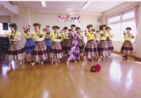 たくさんの女性が、お揃いの衣装とレイを身につけ、室内でフラダンスを踊っている写真