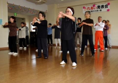 広いホールで、たくさんの男女が正面を向いて並んで立ち、両手を前に組んだポーズをしている写真