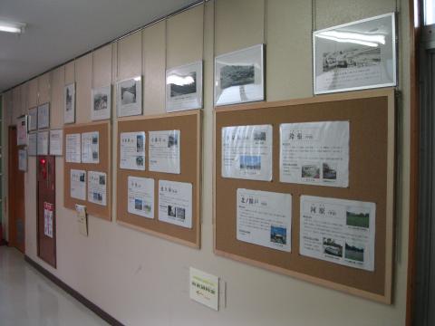 建物内の廊下の壁の掲示板に写真付きの資料が複数貼られ、額に入った写真が複数掛けられて展示されている様子の写真