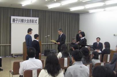 出席者が着席している中、親子川柳大会表彰式の横断幕が貼られたボードの前で、スタンドマイクの前に立った男性が正面に並んで立った父子に向かって賞状を読み上げている様子の写真