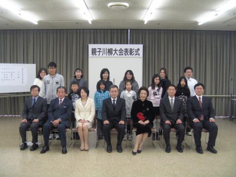 親子川柳大会表彰式の横断幕が貼られたボードの前で、7人のスーツ姿の男性と女性が椅子に座り、その後ろに6組の親子が並んで立って写した記念写真