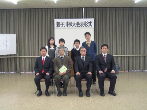 親子川柳大会表彰式の横断幕が貼られたボードの前で、4人のスーツ姿の男性が椅子に座り、その後ろに2組の親子が並んで立って写した記念写真