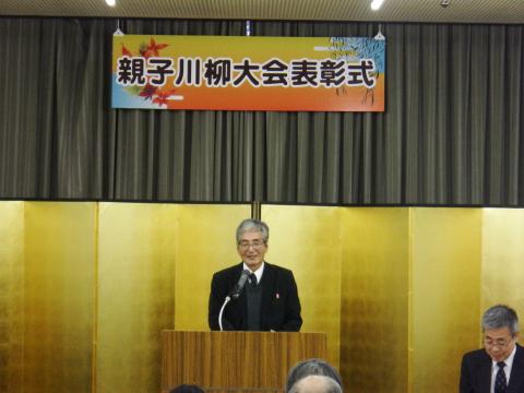 「親子川柳大会表彰式」の横断幕の下に設置した金屏風の前で、演台に立ったスーツ姿の男性が、マイクに向かってにこやかに話をしている様子の写真