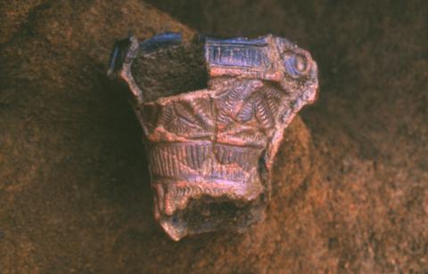 幾何学的な文様が複雑に施され、一部が欠損している土器が土の上に置かれている写真