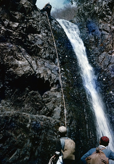 切り立った岩肌を流れ落ちる滝の横で、1人の人物がロープを使って上まで登っており、下で2人の男性がその様子を見守っている写真