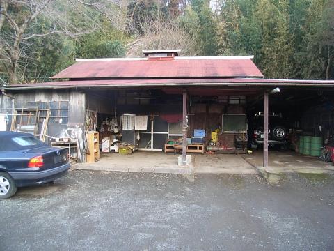 林を背後に建つ、広い軒下の作業スペースがある赤い屋根の平屋の建物の写真