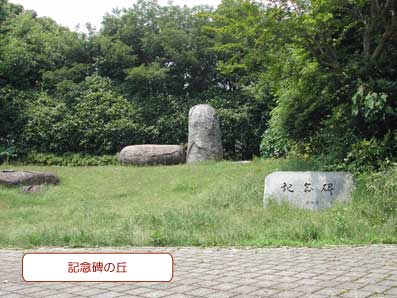 緑の木々に囲まれた芝生広場の中に、縦長の石と横長の石が隣り合って並んだモニュメントがあり、手前に「記念碑」と彫られた石が設置されている写真に「記念碑の丘」の文字が記載された画像