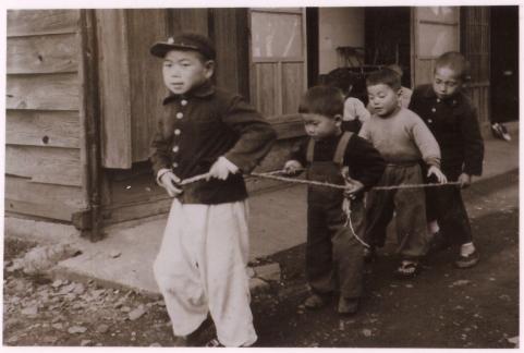 木造の家の前で、数名の子どもたちが輪にした紐の中に入り、電車ごっこをして遊んでいる様子の白黒写真