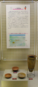 縄文土器と見られる複数の土器が展示台に並べられ、背景の壁には小南遺跡に関する説明文と地図や航空写真が掲示されている写真