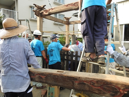 複数の作業者が柵の中で灯籠を建てており、それを周で見ている人たちがいる作業の様子の写真