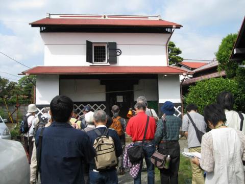 白い壁に2段の赤い屋根が付いた蔵の前に、資料を手にした参加者たちが集まり、見学をしている様子の写真