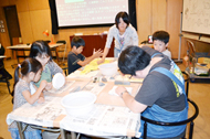 広間のテーブルに集まった親子や子どもたちが、紙や土のような素材を使い、熱心に工作に取り組んでいる様子の写真