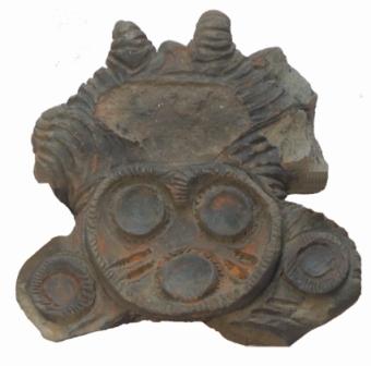 濃い灰色で茶色がかった部分がある、線で表した羽に囲まれた顔の中に、円形の目と口の装飾が施されたみみずく土偶の頭部の写真