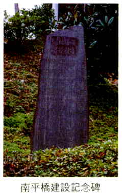 斜面に植えられた低木の中に先が斜めになった長方形の濃いグレーの石碑が立っている写真の下に「南平橋建設記念碑」と書かれた画像