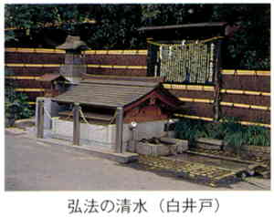竹垣の前に文字が書かれた案内板が設置されており、その前に柵で守られた、石組みと水路があり低い屋根がついた小さな建造物がある写真の下に「弘法の清水（白井戸）」と書かれた画像
