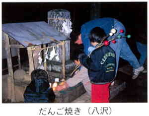 夜の屋外で、石碑の横に建つ道祖神が祭られた小さな祠に子どもたちがお供え物をしている様子の写真に「だんご焼き（八沢）」と書かれた画像