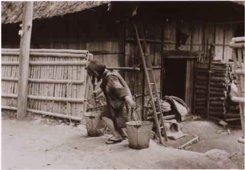 木造藁ぶき屋根の家の前の道で、小さな女の子が左右の手に持った大きなバケツを持ち上げて歩き出そうとしている様子の白黒写真