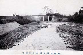 細い川の奥に見えるダムのような構造物の上を機関車が走っている様子をとらえた白黒写真