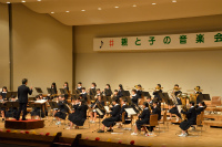 舞台上で黒い制服を着た大勢の生徒たちが、指揮者のもとで管楽器や弦楽器を演奏している写真