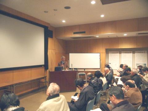 会場で、多くの参加者たちが椅子に座って前方にあるスクリーンと小川さんに注目しており、小川さんが講演台から聴衆に向かって話している様子の写真