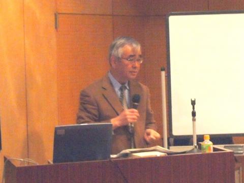 茶色いジャケットとネクタイを着用した小川さんが、演台の前に立ち、マイクを持って話をしている様子の写真