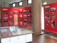 赤い展示パネルに多数の写真が展示されており、その手前のガラスケースの中には複数の資料が展示されている、パネル展の様子の写真